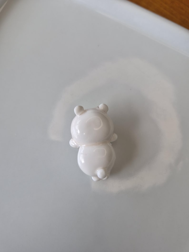 May include: A small, white, glossy figurine of a bear-like character. The figurine has rounded features, including a circular body, small arms, and a pair of ears. The bear is standing upright, with its back to the viewer.