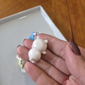 May include: A close-up of a hand holding a small, white, ceramic bear figurine. Other figurines, including a blue animal and a yellow bunny, are visible on a white tray. The figurines have a glossy finish and are likely decorative items.