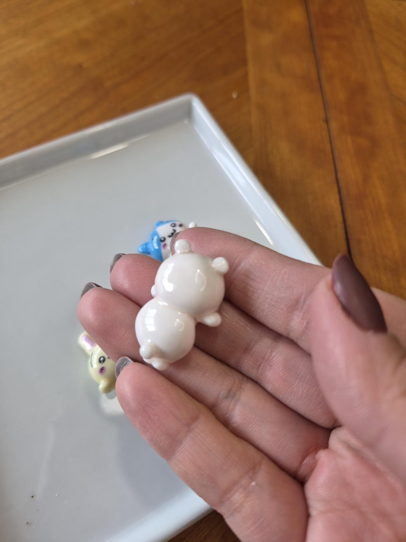 May include: A close-up of a hand holding a small, white, ceramic bear figurine. Other figurines, including a blue animal and a yellow bunny, are visible on a white tray. The figurines have a glossy finish and are likely decorative items.