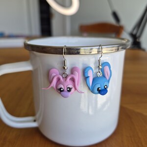 Puede incluir: Pendientes hechos a mano, uno rosa y otro azul, cada uno con un dise&ntilde;o de conejito de dibujos animados. Cuelgan de una taza de esmalte blanco con borde plateado. Las caras de los conejos tienen ojos negros y detalles morados.