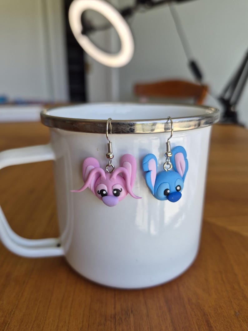 Puede incluir: Pendientes hechos a mano, uno rosa y otro azul, cada uno con un dise&ntilde;o de conejito de dibujos animados. Cuelgan de una taza de esmalte blanco con borde plateado. Las caras de los conejos tienen ojos negros y detalles morados.
