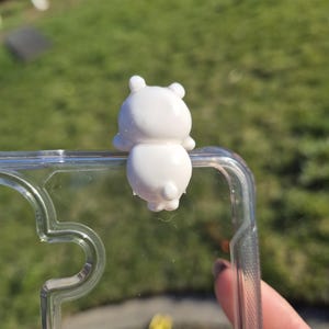 May include: A white, bear-shaped phone charm attached to a clear phone case. The charm has a rounded body, small ears, and a tiny tail. The phone case is transparent, allowing the phone's design to be visible. The background is a blurred green lawn.