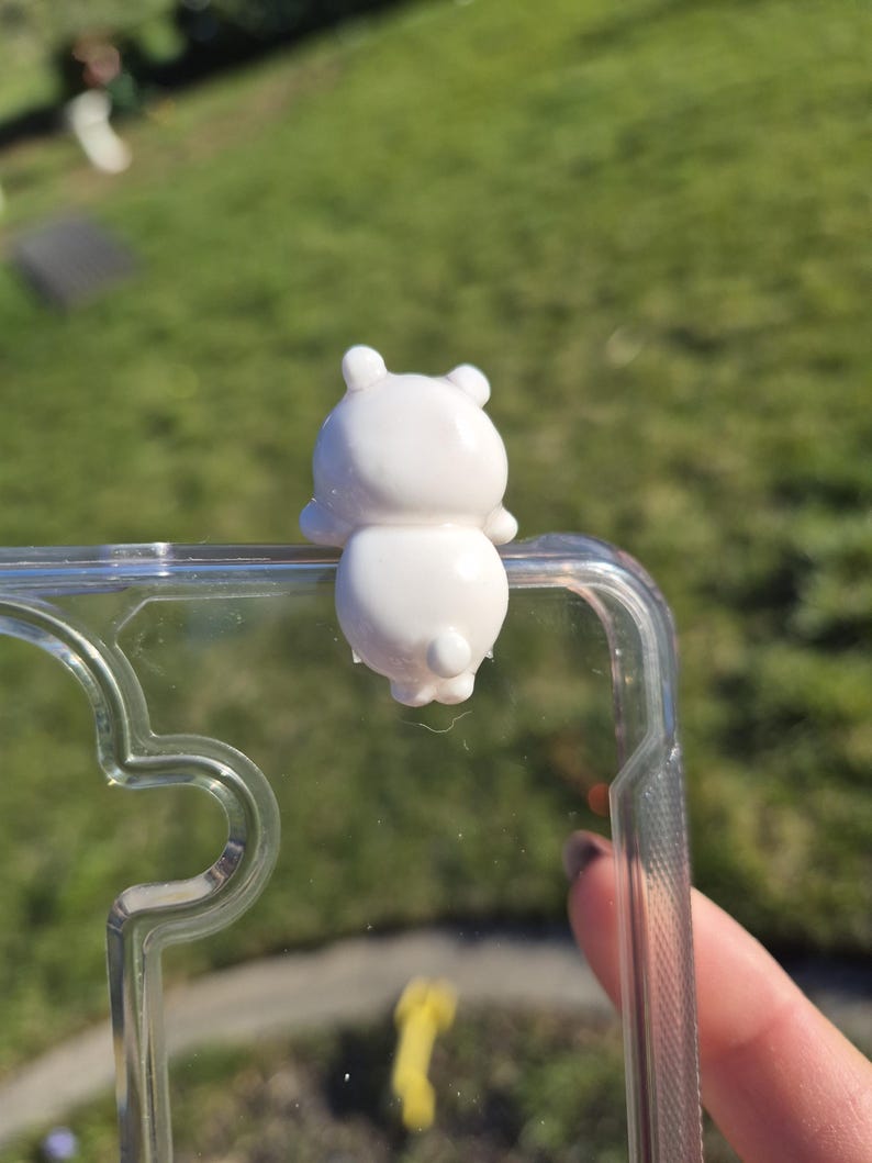 May include: A white, bear-shaped phone charm attached to a clear phone case. The charm has a rounded body, small ears, and a tiny tail. The phone case is transparent, allowing the phone's design to be visible. The background is a blurred green lawn.