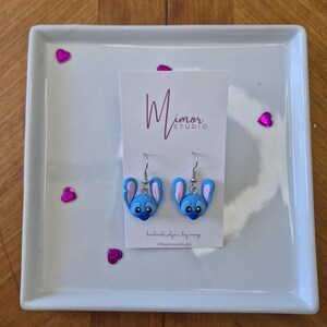 Pu&ograve; includere: Un paio di orecchini in pasta polimerica blu e rosa a forma di testa di personaggio dei cartoni animati, esposti su una piastra quadrata bianca. Gli orecchini hanno ganci argentati e sono presentati su una carta con il testo "Mimor Studio".