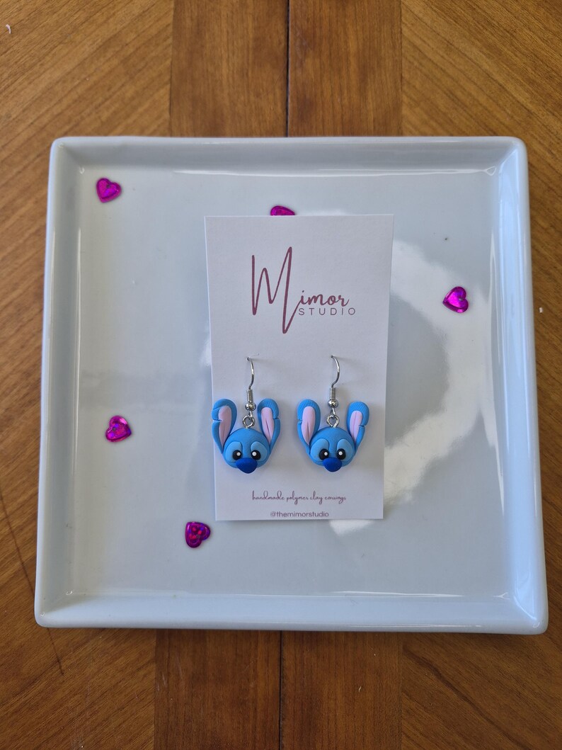 Pu&ograve; includere: Un paio di orecchini in pasta polimerica blu e rosa a forma di testa di personaggio dei cartoni animati, esposti su una piastra quadrata bianca. Gli orecchini hanno ganci argentati e sono presentati su una carta con il testo "Mimor Studio".