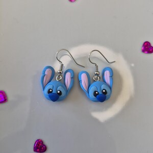 Pu&ograve; includere: Un paio di orecchini blu e rosa a forma di personaggio, con ganci argentati. Gli orecchini presentano un design di personaggio dei cartoni animati con grandi orecchie e un naso blu scuro. Su sfondo bianco, con coriandoli rosa a forma di cuore.