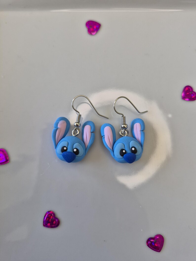 Pu&ograve; includere: Un paio di orecchini blu e rosa a forma di personaggio, con ganci argentati. Gli orecchini presentano un design di personaggio dei cartoni animati con grandi orecchie e un naso blu scuro. Su sfondo bianco, con coriandoli rosa a forma di cuore.