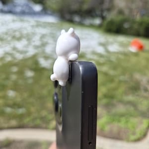 May include: A white, cartoon-style bunny phone grip attached to a black smartphone. The bunny is small and has a rounded body with two upright ears. The phone is held against a blurred green and white outdoor background.