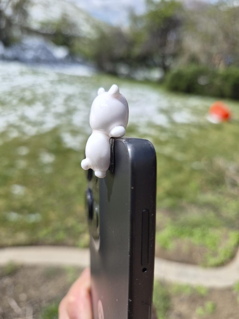 May include: A white, cartoon-style bunny phone grip attached to a black smartphone. The bunny is small and has a rounded body with two upright ears. The phone is held against a blurred green and white outdoor background.