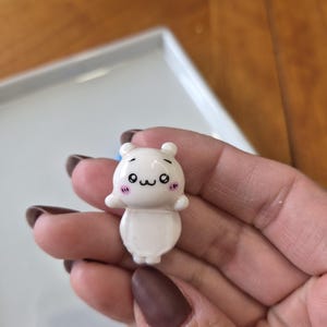 May include: A small, white figurine of a cartoon bear with a cute expression. The bear has small ears, black eyes, and pink cheeks. It is held in a hand against a blurred background.