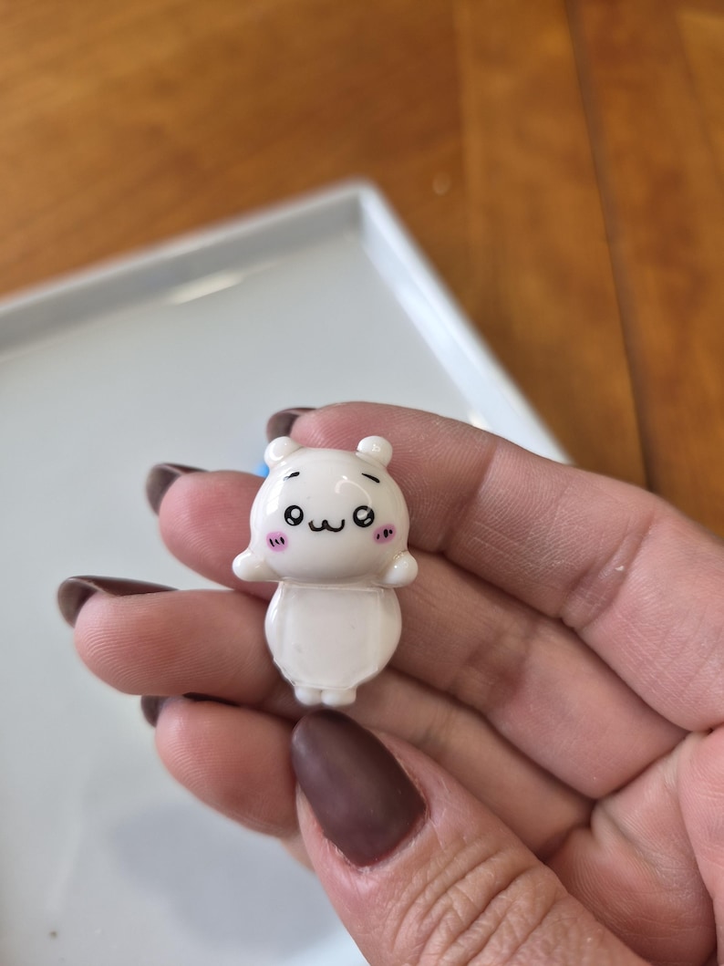 May include: A small, white figurine of a cartoon bear with a cute expression. The bear has small ears, black eyes, and pink cheeks. It is held in a hand against a blurred background.