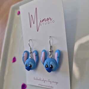 Pu&ograve; includere: Un paio di orecchini in argilla polimerica blu a forma di personaggio dei cartoni animati con grandi orecchie e un naso blu scuro. Gli orecchini sono su una carta bianca con il testo "Mimor Studio" e "handmade polymer clay earrings".