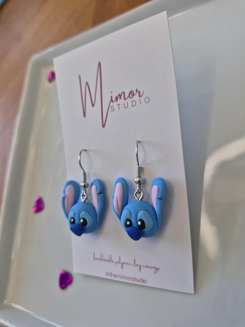 Pu&ograve; includere: Un paio di orecchini in argilla polimerica blu a forma di personaggio dei cartoni animati con grandi orecchie e un naso blu scuro. Gli orecchini sono su una carta bianca con il testo "Mimor Studio" e "handmade polymer clay earrings".