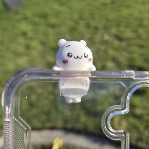 May include: A white, cartoon-style bear figure with black eyes and a small mouth, attached to a clear phone case. The bear has a cute expression. The background is blurred green grass.