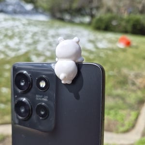 May include: A small, white bear figurine perched on the edge of a black smartphone. The phone has multiple camera lenses and the text "50MP OIS QUAD PIXEL". The background is a blurred outdoor scene with green grass and trees.