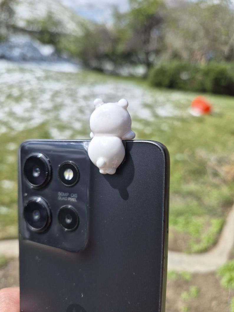 May include: A small, white bear figurine perched on the edge of a black smartphone. The phone has multiple camera lenses and the text "50MP OIS QUAD PIXEL". The background is a blurred outdoor scene with green grass and trees.