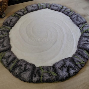 May include: A circular rug featuring a central white area with a subtle spiral texture, surrounded by a border of dark gray, textured blocks that resemble stones. Small green tufts are interspersed between the gray stone-like segments.