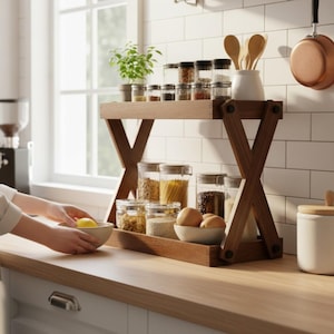 May include: A two-tiered wooden spice rack with a dark brown finish. The top shelf holds small glass jars of spices, while the bottom shelf displays clear jars of pasta, grains, and eggs. A small potted plant and wooden spoons are also visible.