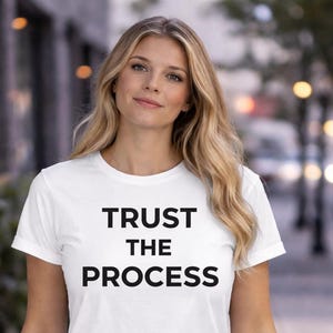 May include: White t-shirt with the text "TRUST THE PROCESS" in black, centered on the front. The shirt has short sleeves and a crew neck. The model has blonde hair.