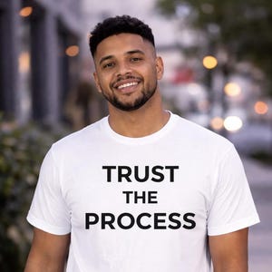 May include: White t-shirt with the text "TRUST THE PROCESS" in black block letters. The shirt is being worn by a person with a beard and short, dark hair. The background is blurred, suggesting an outdoor setting.