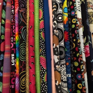 Peut inclure: Une collection de rouleaux de tissu colorés avec divers motifs. Les motifs comprennent des cœurs, des motifs tie-dye, des tranches de pastèque et des visages de chiens. Les tissus sont dans des tons de rouge, noir, rose, vert et bleu.