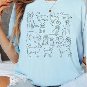 Peut inclure: T-shirt bleu clair orné d'illustrations de chiens en noir. Le motif présente différentes races, comme un Golden Retriever, un Caniche et un Teckel. Un style décontracté et confortable.
