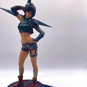 May include: A detailed figurine of a woman in a futuristic outfit. She wears a dark green top, black shorts, and brown boots. The figure has a black helmet with sharp, wing-like extensions. The base is a stylized shape with red and blue accents.