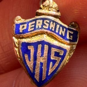 May include: A gold-toned metal shield-shaped pendant featuring blue enamel inlay. The top banner displays the text PERSHING in gold lettering, while the lower section of the shield features the gold letters JHS centered against a blue background.