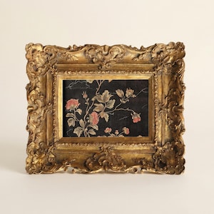 May include: A rectangular picture frame with an ornate, embossed gold finish. The frame surrounds a dark black fabric insert featuring a printed pattern of green leaves and pink flower blossoms with thin, light-colored stems and branches.