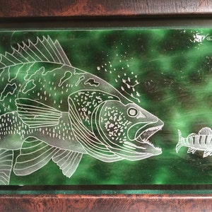May include: A framed etched glass artwork depicting a largemouth bass with its mouth open, chasing a smaller fish. The background is a green and white marbled design. The artwork is signed "© 2015".