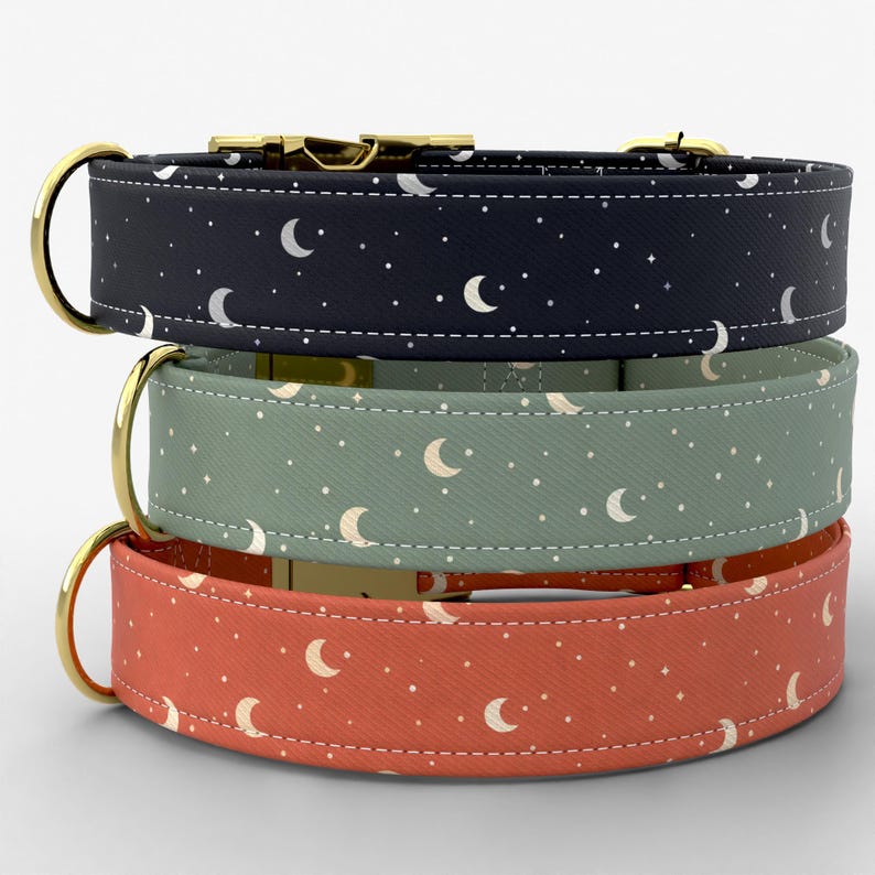 May include: A stack of three pet collars featuring a celestial pattern of white crescent moons and stars. The top collar is dark navy, the middle is sage green, and the bottom is terracotta orange. Each collar has gold-toned metal hardware.