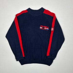 May include: A navy blue long-sleeve knit jumper featuring red stripes running down the shoulders and sleeves. The chest displays the red text FUBU INTERNATIONAL above small flags of the United States, Canada, and the Netherlands, with a flag patch.