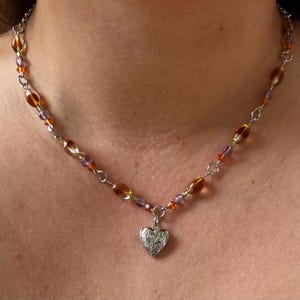 May include: A beaded necklace with a silver chain and a heart-shaped locket pendant. The necklace features a mix of amber, purple, and green beads. The locket has a floral design.