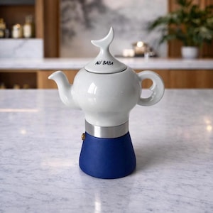 Puede incluir: Una tetera de cerámica blanca y azul con un diseño único. La tetera tiene un cuerpo blanco, una base azul y una banda plateada. La tapa está adornada con un asa en forma de aleta, y el texto "ALI BABA" está impreso en la tapa.