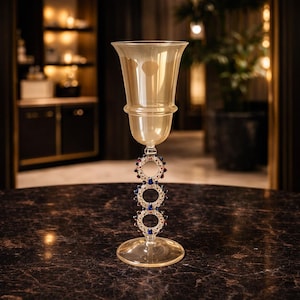 Peut inclure: Un gobelet en verre orné, avec un bord évasé et une tige décorative. La tige présente trois motifs circulaires ornés de pierres précieuses rouges et bleues. Le verre est de couleur ambre clair et repose sur une base ronde.