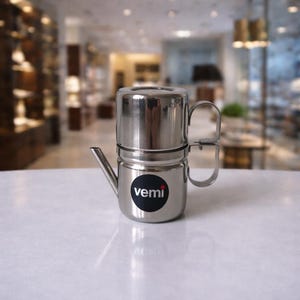 Op de afbeelding: Een roestvrijstalen koffiezetapparaat met een cilindrisch ontwerp, met een tuit en twee handvatten. De koffiezetter heeft een zwart rond label met het woord "vemi" in het wit. Het product staat op een wit oppervlak.