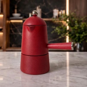 Pode incluir: Uma cafeteira italiana vermelha com uma alça prateada e uma bola prateada no topo. A cafeteira é feita de metal e tem um acabamento texturizado. A cafeteira está em uma bancada de mármore.