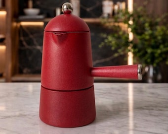 May include: A red stovetop espresso maker with a silver handle and a silver ball on top. The coffee maker is made of metal and has a textured finish. The espresso maker is on a marble countertop.