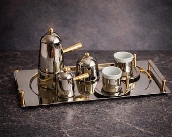 Könnte beinhalten: Ein poliertes silbernes Kaffeeservice mit goldenen Akzenten, bestehend aus einer Kaffeekanne, einem Sahnekännchen, einer Zuckerdose und zwei Tassen, alles auf einem rechteckigen Tablett. Das Set hat ein modernes Design, ideal für Kaffee oder Tee.