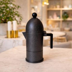 Könnte beinhalten: Ein dunkelgrauer Espressokocher mit schwarzem Griff und kleinem schwarzem Knopf. Der Kaffeezubereiter ist zylindrisch mit Ausguss und steht auf einer Marmoroberfläche. Der Hintergrund ist unscharf, zeigt eine Küchenumgebung.