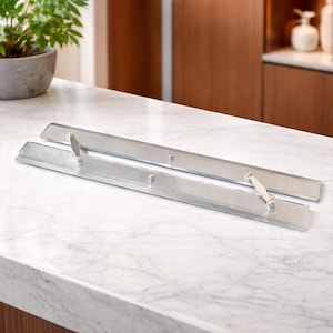 Puede incluir: Dos tiradores de armario rectangulares plateados con asas decorativas se exhiben sobre una encimera de mármol blanco. Los tiradores tienen un diseño elegante y moderno, y probablemente se utilizan para cajones o puertas de armarios.