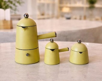May include: A set of three yellow coffee pots with silver accents. The largest pot has a two-part design with a handle. The smaller pots have a rounded shape with a lid and a handle. The pots are on a light-colored surface.