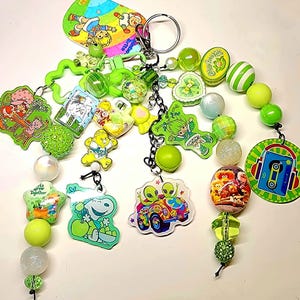 May include: A collection of colorful keychains and beaded accessories. The keychains feature cartoon characters and designs, including aliens, Snoopy, and Strawberry Shortcake. The beads are in shades of green, white, and iridescent colors.