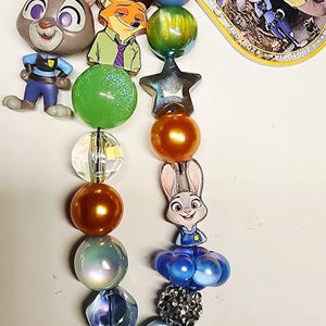 Op de afbeelding: Een Zootopia-sleutelhanger met een goudkleurige karabijnhaak, een ronde Zootopia-logobedel en twee strengen met kleurrijke kralen. De kralen hebben diverse vormen, maten en afwerkingen, afgewisseld met bedels van Judy Hopps en Nick Wilde.