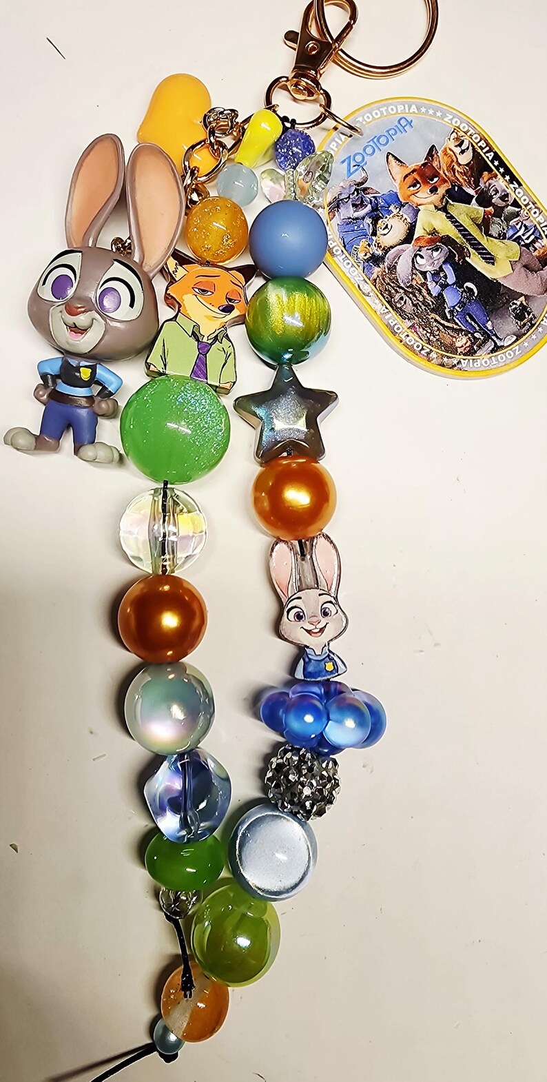 Op de afbeelding: Een Zootopia-sleutelhanger met een goudkleurige karabijnhaak, een ronde Zootopia-logobedel en twee strengen met kleurrijke kralen. De kralen hebben diverse vormen, maten en afwerkingen, afgewisseld met bedels van Judy Hopps en Nick Wilde.
