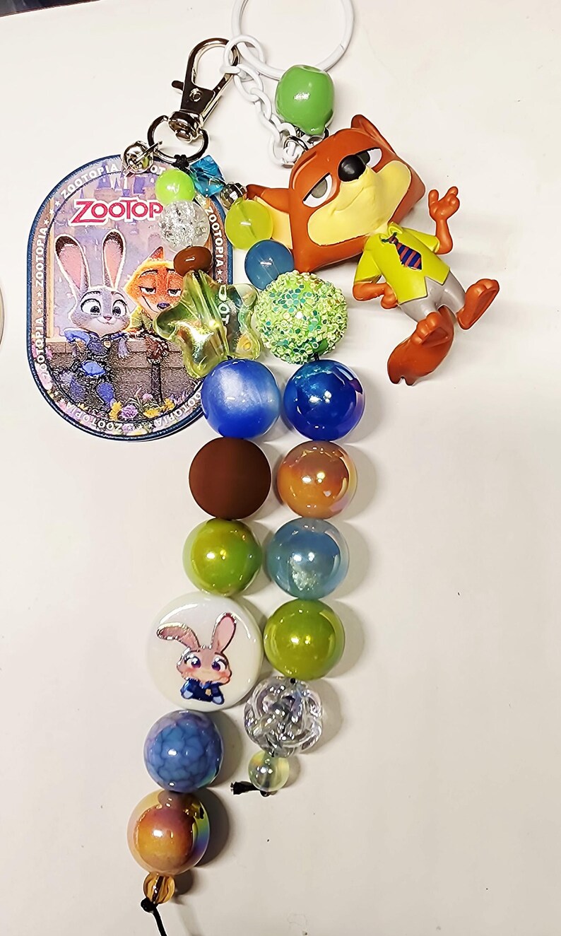 Op de afbeelding: Een Zootopia-sleutelhanger met een metalen sluiting, een ronde bedel met Judy Hopps en Nick Wilde, een Nick Wilde-figuurtje en twee strengen veelkleurige kralen, waaronder groene, blauwe, bruine en iriserende bollen met een Judy Hopps-kraal.