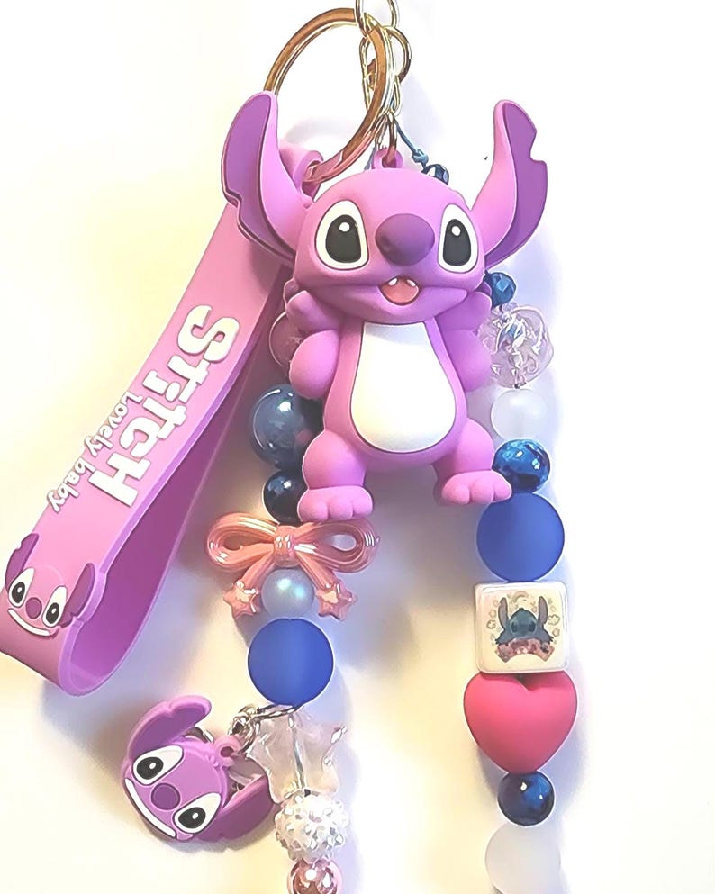 May include: A pink Stitch keychain with a gold-colored ring. The keychain features a Stitch figure, a pink strap with "Stitch lovely baby" text, and decorative beads in pink, blue, and white. A pink bow and a heart-shaped bead are also included.