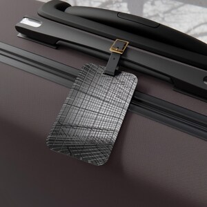 May include: A close-up of a dark gray suitcase with a black handle and a luggage tag. The luggage tag is rectangular with a textured, grid-like pattern. The tag is attached with a black strap and a gold-colored buckle.