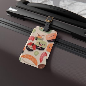 May include: A luggage tag featuring a repeating pattern of smiling sushi characters in various colors. The tag has a beige background and a black strap with a gold-colored buckle. The tag is rectangular and made of a glossy material.