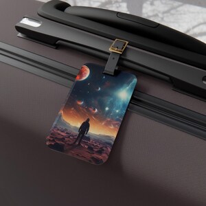 Puede incluir: Una etiqueta de equipaje rectangular con una vibrante escena espacial con una silueta en un planeta rocoso. La etiqueta tiene una correa negra con una hebilla dorada, sujeta a una maleta gris oscuro.